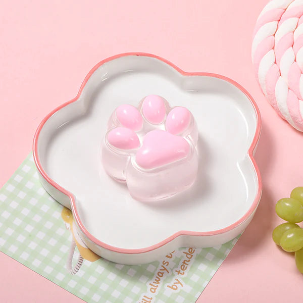 White scalloped dish with a pink rim holding a glossy pink paw-shaped item on a pastel pink background